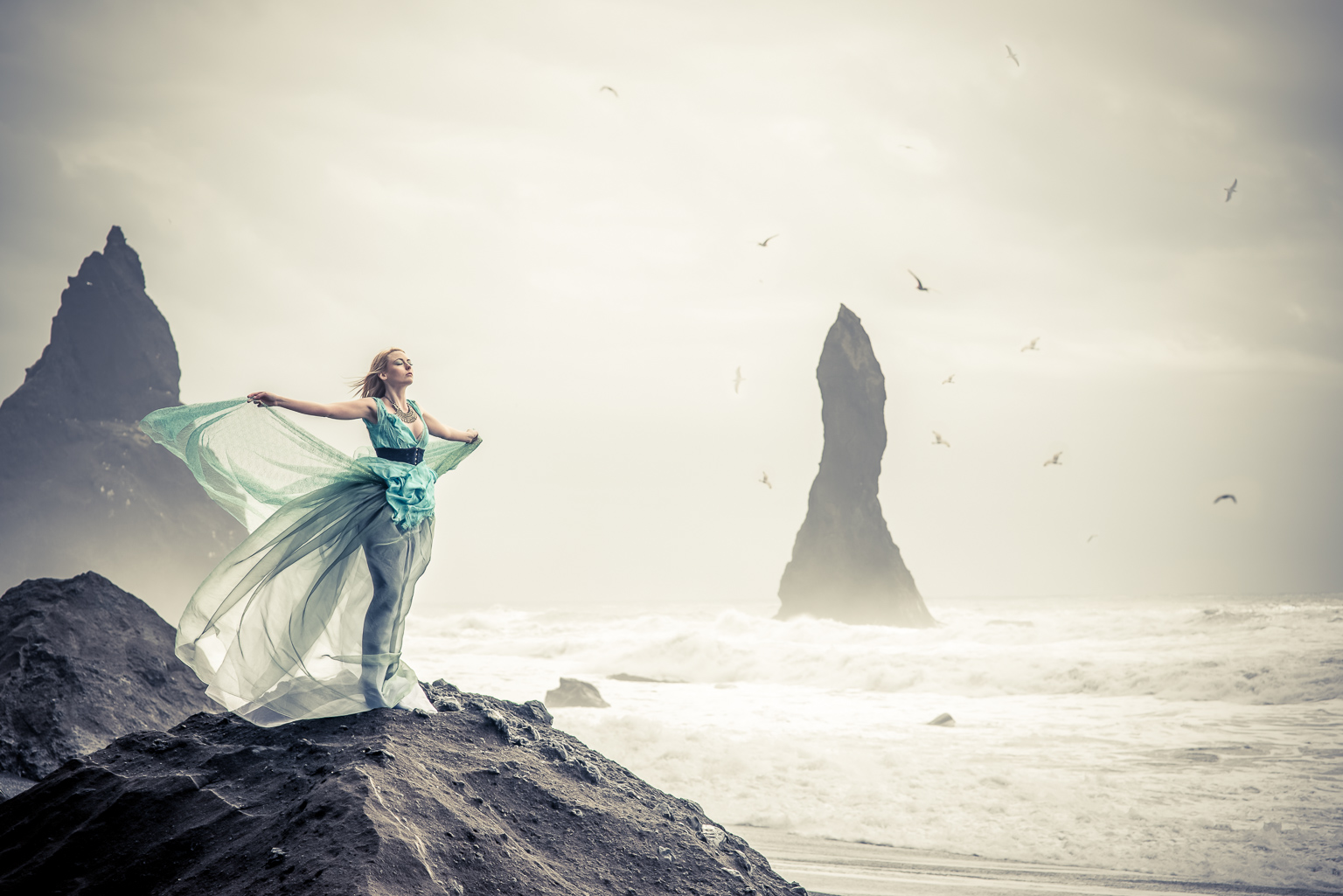 A model in a sea green floaty dress standing on a rock by an angry sea in Iceland. Seagulls flying in the distance