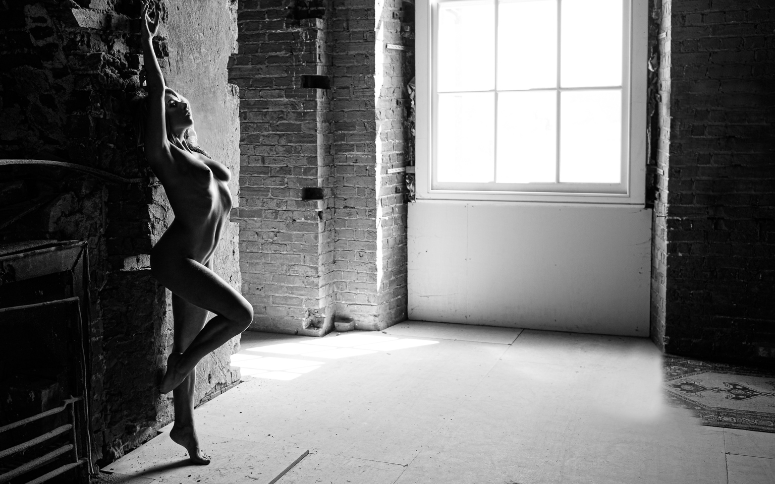 art nude model stretching up against a brick fireplace in deep shadow with curves of body highlighted by light from a large window