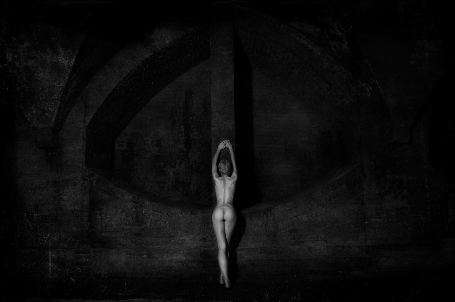 art nude shot of model stretched upright facing a brick structure in a church cellar. black and white by ian mears