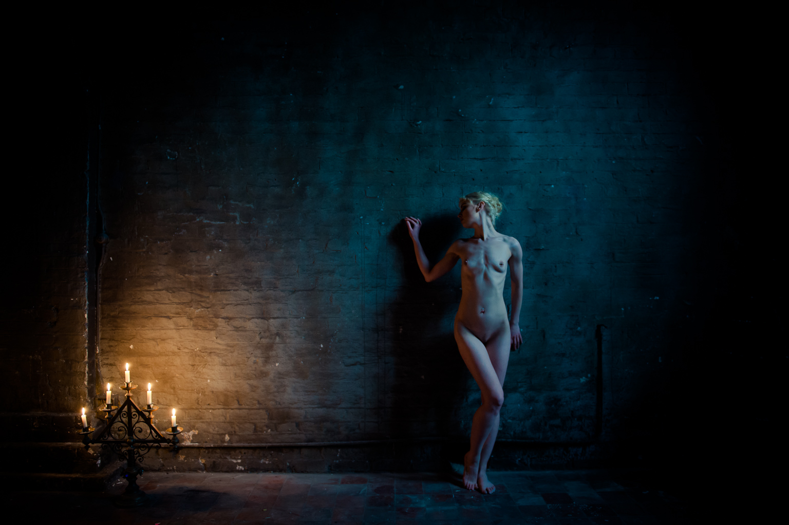an art nude model leaning, face on to camera, against a brick wall. Cool blue light from a window to the right and warm candlelight from the left. taken in a church in London by Ian Mears